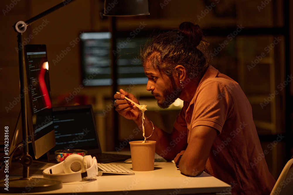 Young programmer examining codes on computer monitor and having lunch while working till late night in office Young programmer examining codes on computer monitor and having lunch while working till late night in office