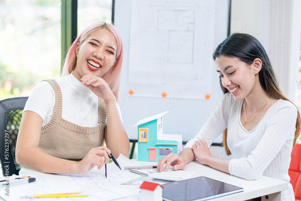 Fototapeta premium Concept of engineering consulting, Two female engineers discussing about model of building together; Two female architects are studying blueprint of building house.