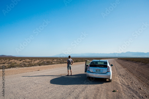 ARIZONA, USA. A man urinates on an empty dirt road leading into a vast desert. His hybrid car waits next to him. photo