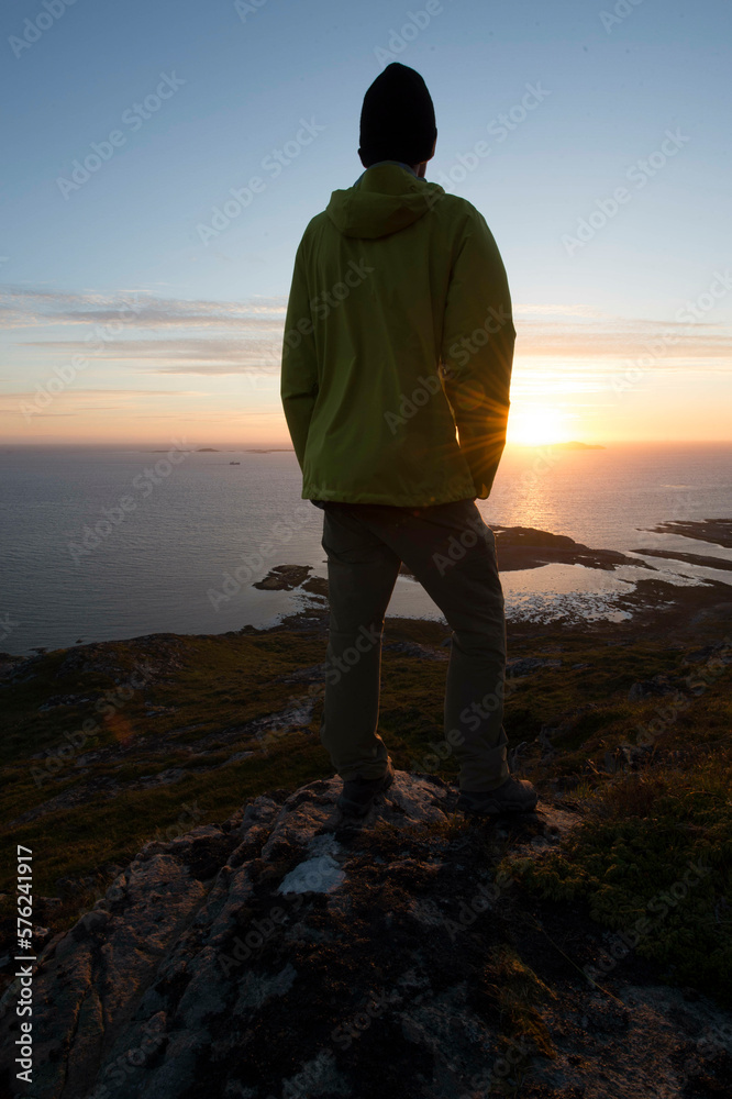 A young male hike stands on top of a mountain on the island of Linesøya and looks into the midnight sun A young male hike stands on top of a mountain on the island of Linesøya and looks into the midnight sun