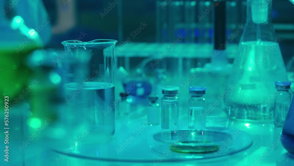 Scientist pours powder into a clear liquid in a flask, it dissolves, turning the liquid green. A woman stirs the solution with a glass pipette. Vials with vaccines and test tubes. Blue light. Close up Scientist pours powder into a clear liquid in a flask, it dissolves, turning the liquid green. A woman stirs the solution with a glass pipette. Vials with vaccines and test tubes. Blue light. Close up