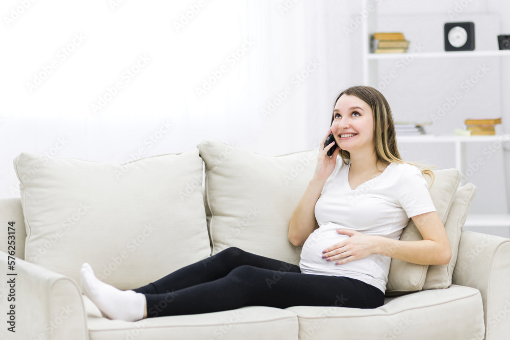 Smiling pregnant woman talking over the phone on white sofa.