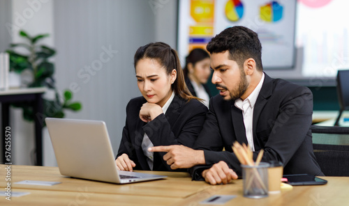 Millennial Asian Indian professional successful bearded male businessman mentor pointing coaching helping female businesswoman employee colleague in formal business suit working via laptop computer