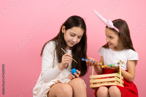 Two sister, one of them wearing bunny ears and keeping a basket with eggs on her knees while another giel painting the egg. photo