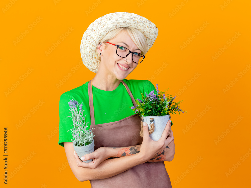 young gardener woman with short hair in apron and hat holding potted plants smiling with happy face looking at camera standing over orange background young gardener woman with short hair in apron and hat holding potted plants smiling with happy face looking at camera standing over orange background
