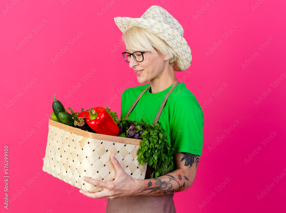 young gardener woman with short hair in apron and hat holding crate full of vegetables looking at them smiling standing over pink background