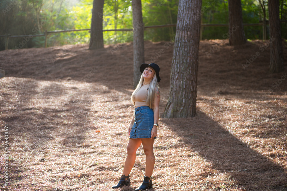 Beautiful blonde woman with straight hair is dressed in brown top and short denim skirt and hat. The woman is happy and walks among the pine trees in the forest. Sun rays enter from behind. Beautiful blonde woman with straight hair is dressed in brown top and short denim skirt and hat. The woman is happy and walks among the pine trees in the forest. Sun rays enter from behind.
