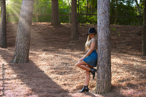 Beautiful blonde woman with straight hair is dressed in brown top and short denim skirt and hat. The woman is happy and leans against a tree in the forest. The sun s rays are coming in from the side.