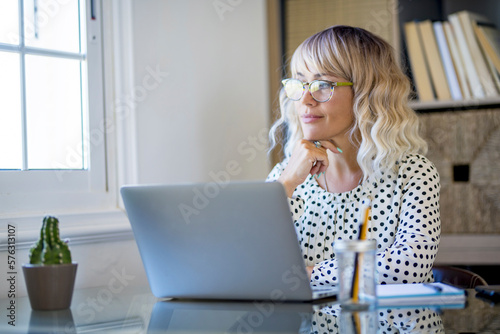 Thoughtful serene business woman looking away thinking solving problem at work, happy smiling young middle age woman relax lost in thought reflecting sit with laptop. Lifestyle of businesswoman