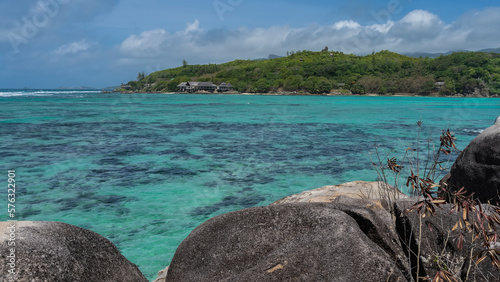 Corals on the bottom are visible through the clear turquoise water of the ocean. Granite boulders in the foreground. In the distance, on the shore of the green island-the villas of the hotel.