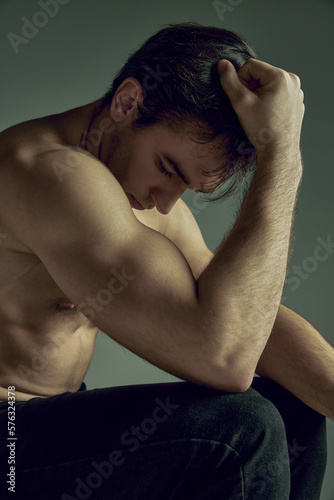 Thoughtful young man with muscular relief body, strong hands shape sitting, posing shirtless in black pants on green studio background. Concept of man's beauty, sportive and healthy lifestyle, fashion photo