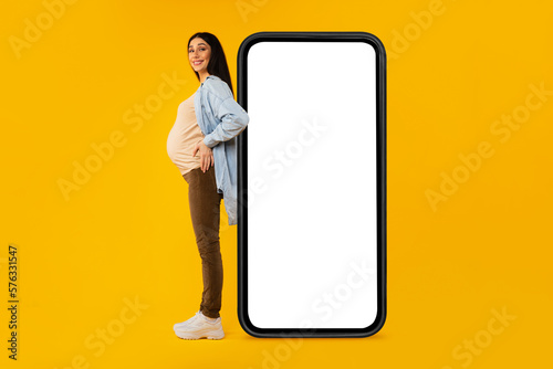 Excited pregnant lady posing near huge smartphone with blank screen over yellow background, full length, mockup © Prostock-studio