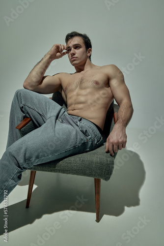 Muscular body. Portrait of young handsome man sitting on chair, posing shirtless in pants over pale green studio background. Concept of man's beauty, sportive and healthy lifestyle, fashion photo