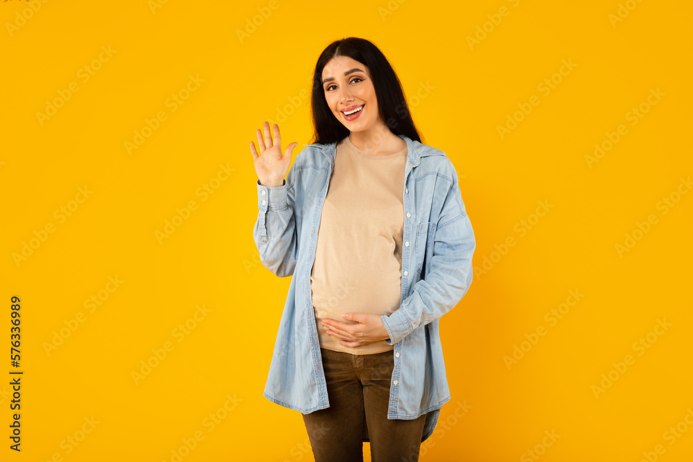 Beautiful expectant woman touching pregnant belly and waving saying hello and smiling at camera, yellow background Beautiful expectant woman touching pregnant belly and waving saying hello and smiling at camera, yellow background