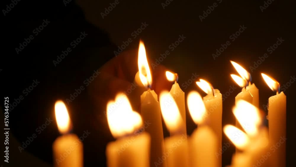 Long white religious candles, flames burning at night in the church. A womans hand lights and puts a candle to others Long white religious candles, flames burning at night in the church. A womans hand lights and puts a candle to others
