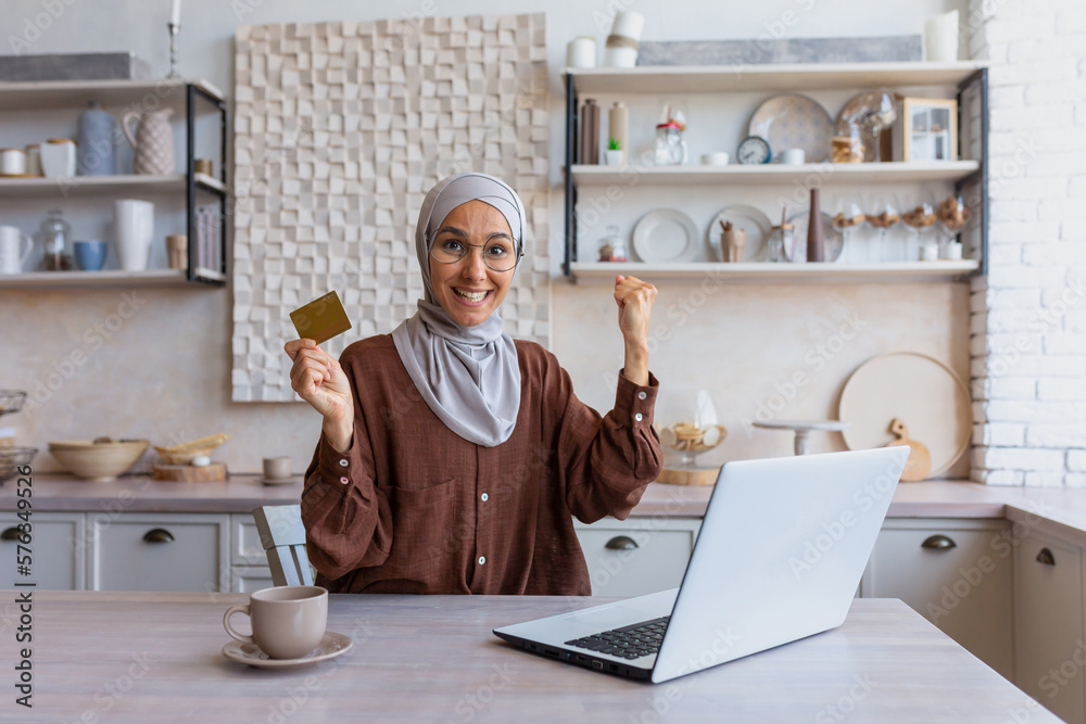 Happy young beautiful Muslim woman in hijab sitting at laptop at home at table, holding and showing credit card in hand, smiling at camera, showing yes hand gesture. Happy young beautiful Muslim woman in hijab sitting at laptop at home at table, holding and showing credit card in hand, smiling at camera, showing yes hand gesture.