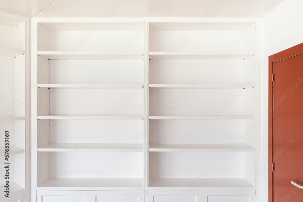 Large wooden closed mahogany door with a brass handle for opening. Along the wall is a white open cabinet with shelves for books and interior items for coziness and comfort. The floor is parquet.