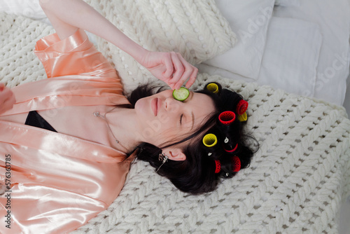 A woman in bathrobe lying on a bed with hair wrapped in curlers and cucumber slices on her eyes. Relaxing on bed wearing face pack and roller curls on hair. At home spa day. Self care and relaxation.