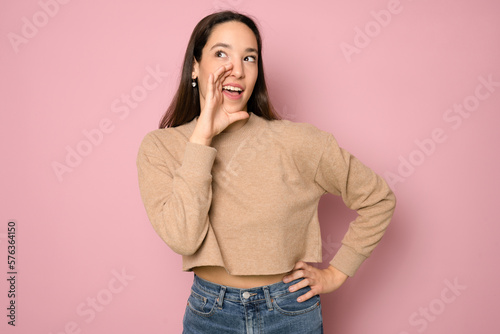 Portrait of a young pretty woman telling secret information, holding her hand near mouth and standing against pink background