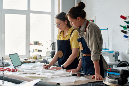 Experienced female tailor putting numbers on sewing paper patterns in atelier while standing by workplace and teaching her subordinate