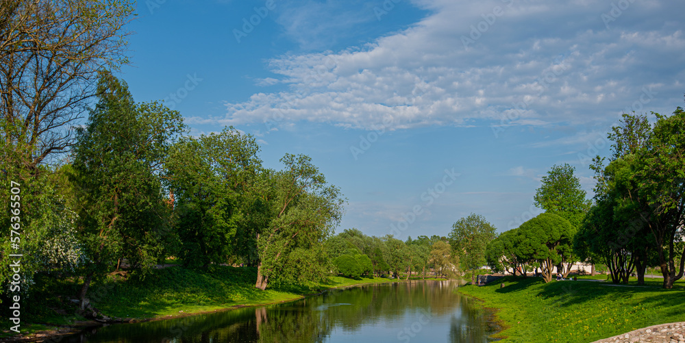 Fototapeta premium Beautiful colorful summer or spring natural landscape with a lake in a park surrounded by green foliage of trees in sunlight and a stone path in the foreground.