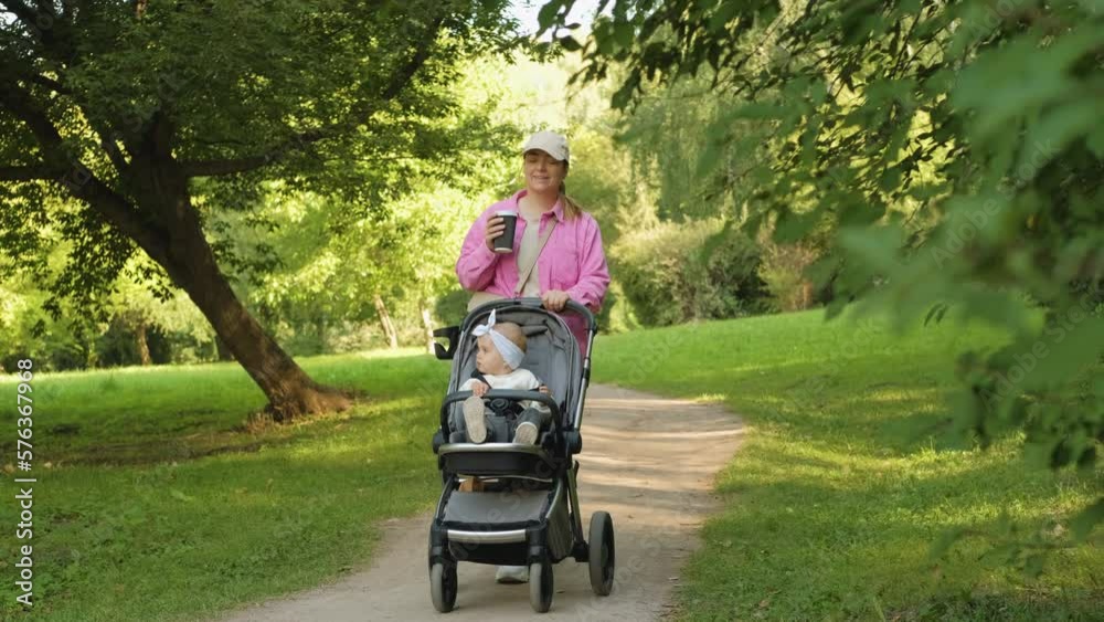 Happy mother drinks coffee while walking in the summer park with her young daughter in a baby carriage outdoors