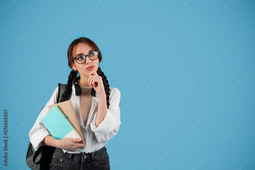 Pensive smiling teenage european girl with pigtails in glasses with backpack hold notebooks, looks up at free space