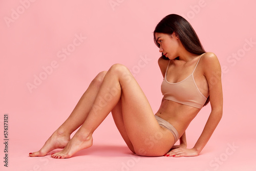 Beautiful young brunette girl with slim body, slender legs posing in cotton underwear over pink studio background. Concept of natural beauty, fitness, diet, body care, sport and health