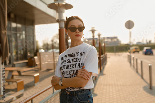 Trendy young woman with dark hairstyle and red lips posing outside. Brunette woman in white t-shirt and glasses looks away outdoors in sunset