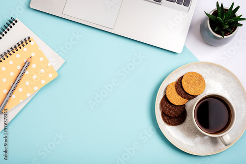 A cup of coffee with cookies, laptop computer, notepads and succulent plant on blue background. Cozy workplace, work from home concept. Top view, flat lay, copy space