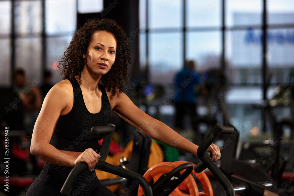 Naklejka premium Positive smiling african american woman, exercising on an exercise bike in the gym, dark background, cardio training on an exercise bike, keep yourself in good shape.