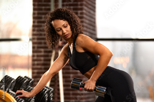 African American woman training with dumbbells in the hall in front of the mirror, against large windows, bright hall, positive view of the woman ternating the muscles of the hands