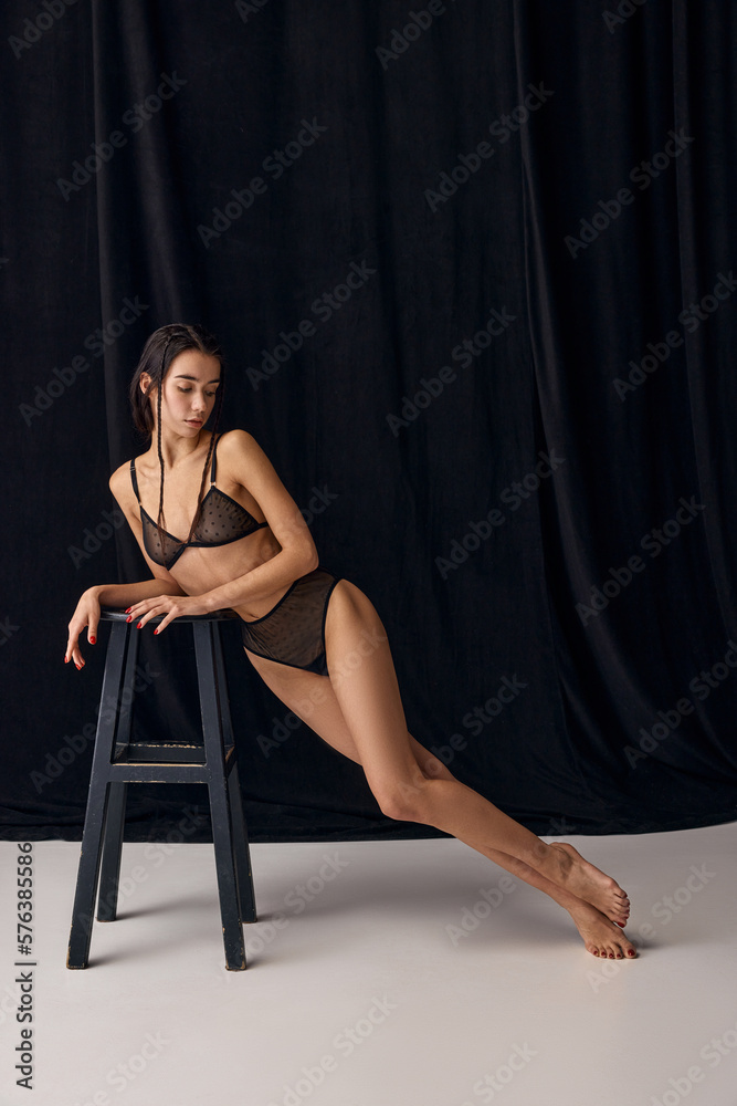Sensuality. Portrait of beautiful young slim girl in underwear leaning on chair, posing over black studio background. Concept of natural beauty, tenderness, femininity, body care and fitness