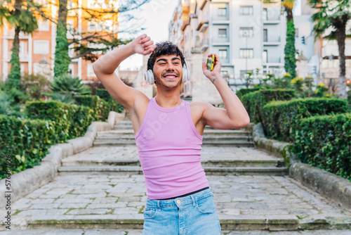Happy young Hispanic male in pink tank top and jeans smiling and looking away while listening to music in wireless headphones with smartphone and dancing with raised arms on street