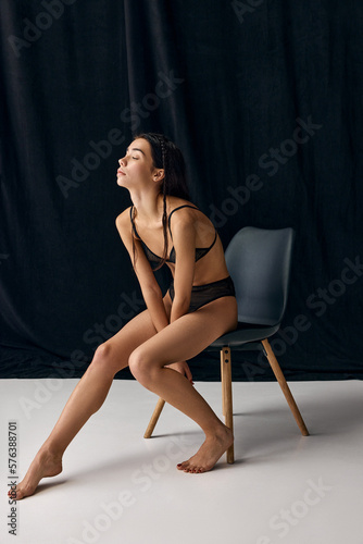 Relaxation. Portrait of beautiful young slim girl in underwear posing on chair over black studio background. Concept of natural beauty, tenderness, femininity, body care and fitness