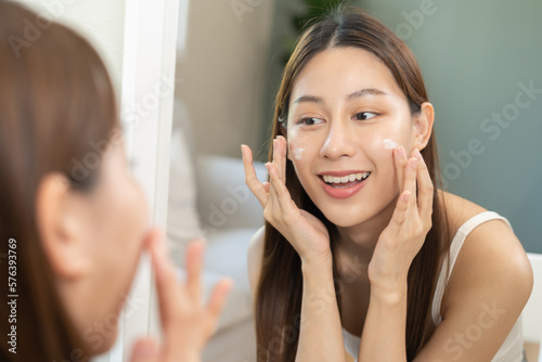 Konstfotografi Fresh healthy skin, beautiful smile of asian young woman, girl looking at mirror, applying moisturizer on her face, putting cream treatment before makeup cosmetic routine at home