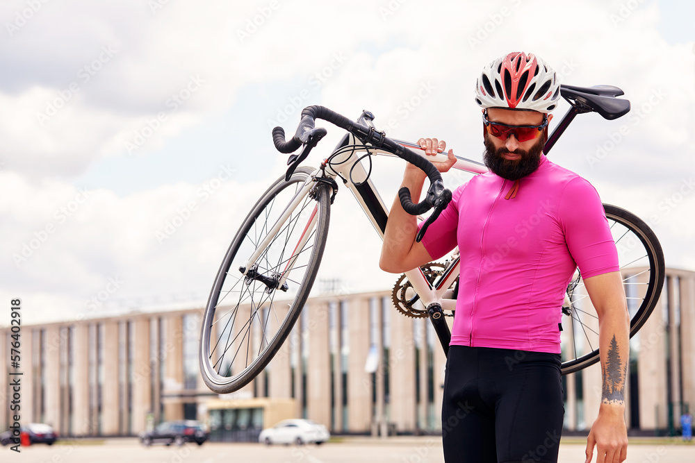 Bearded male cyclist in glasses and a helmet looks at the camera with a bicycle in his hand against the blue sky, lifestyle, cardio workout Bearded male cyclist in glasses and a helmet looks at the camera with a bicycle in his hand against the blue sky, lifestyle, cardio workout