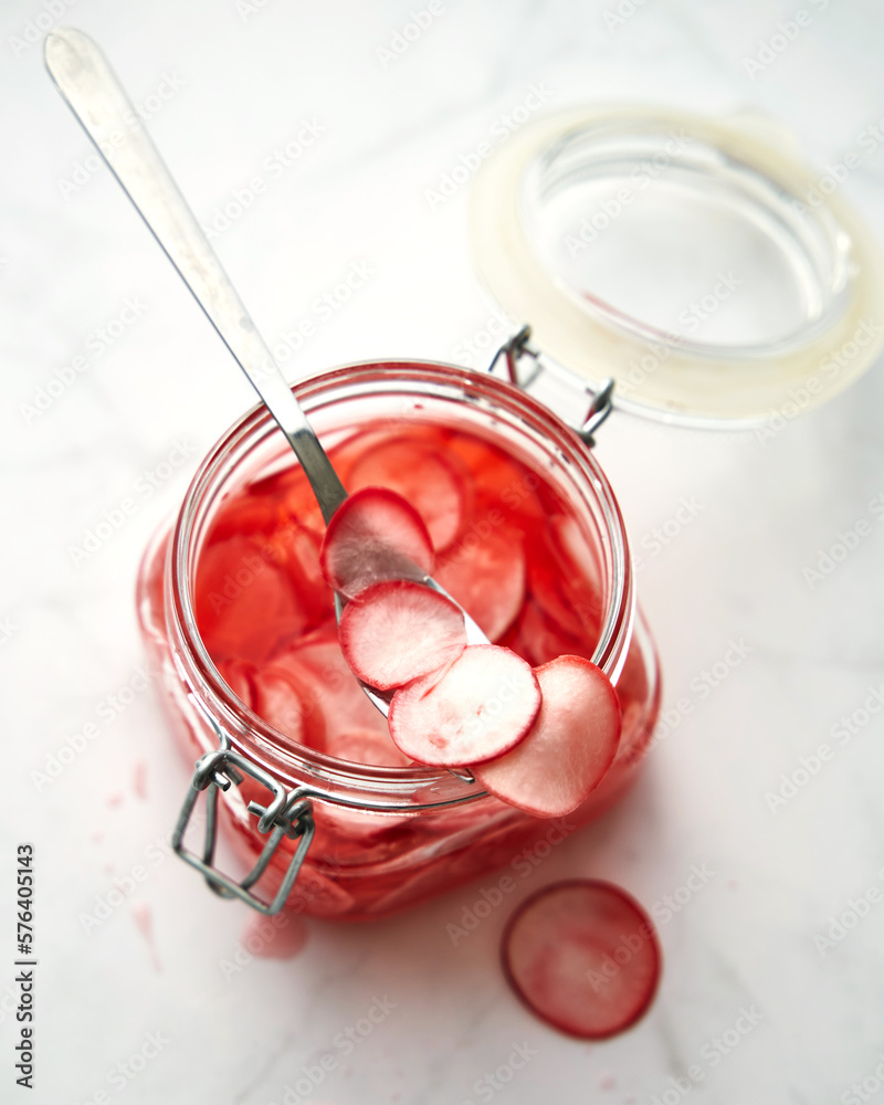 Jar of pickled thinly sliced pink radishes. A spoon is sticking out of the jar. White background. Korean dish.