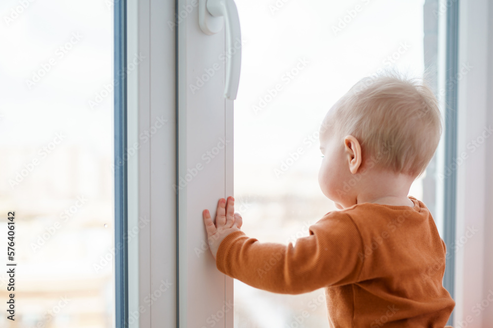 BabyToddler is standing with his hand on the window and looking at the street.