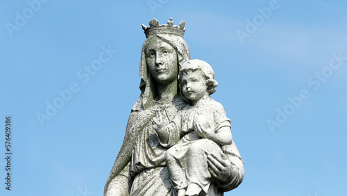 1920s statue of the virgin Mary with baby Jesus in cemetery. Worn, weathered and covered in moss against a blue sky. © John Hanson Pye