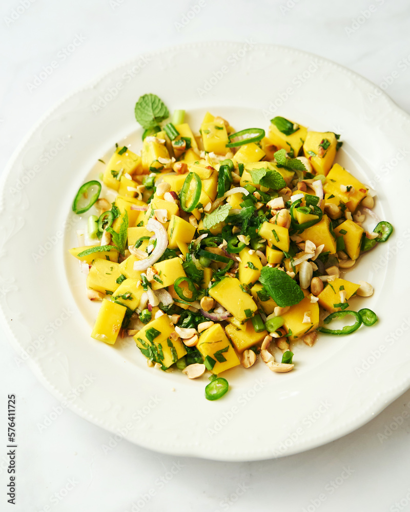 Thai mango salad in the white plate on white background. Salad from mango with peanuts, chili and fish sauce decorated with mint leaves. Thai mango salad in the white plate on white background. Salad from mango with peanuts, chili and fish sauce decorated with mint leaves.