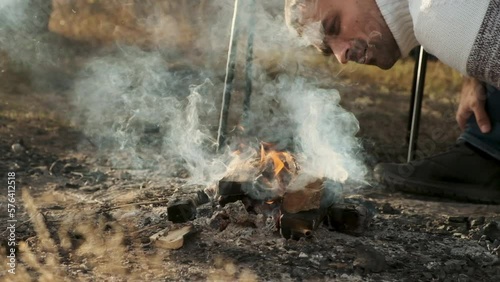 Tourist route through autumn forest. An adult male traveler lights campfire in camp. man blows on flame to kindle it more. close-up. picnic in forest