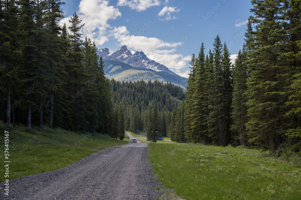 The landscape of the Rocky Mountains. Rural road through the forest - mountains, conifers, saturated blue sky with clouds in the background