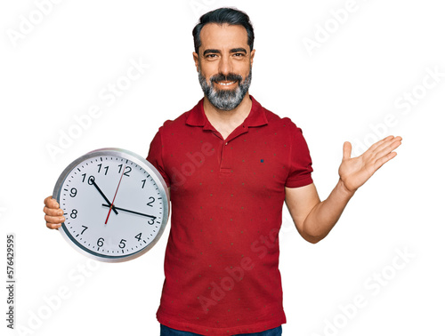 Middle aged man with beard holding big clock celebrating victory with happy smile and winner expression with raised hands
