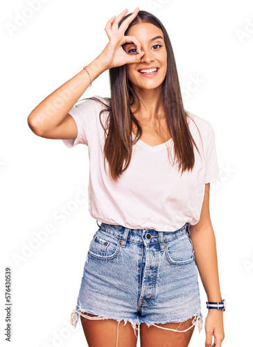 Young hispanic woman wearing casual white tshirt doing ok gesture with hand smiling, eye looking through fingers with happy face.