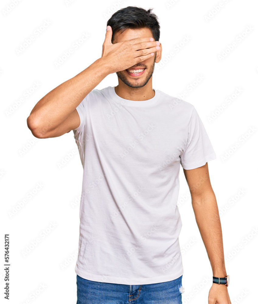 Young handsome man wearing casual white tshirt smiling and laughing with hand on face covering eyes for surprise. blind concept. Young handsome man wearing casual white tshirt smiling and laughing with hand on face covering eyes for surprise. blind concept.