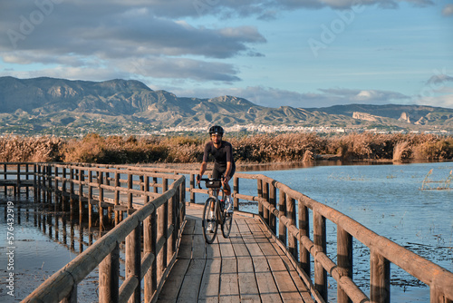 Fototapeta Naklejka Na Ścianę i Meble -  A man is riding over a lake with a background of mountains.
Male cyclist is riding a gravel bike on a wooden bridge over a lake.
Elche, Alicante, Spain