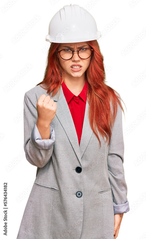 Young redhead woman wearing architect hardhat angry and mad raising fist frustrated and furious while shouting with anger. rage and aggressive concept.