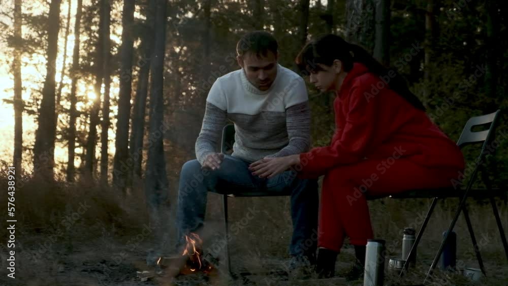 Portrait family sitting by campfire on an autumn evening. Adult woman with man is resting in camping near tent. people at sunset are sitting near campfire drinking tea chatting and warming hands Portrait family sitting by campfire on an autumn evening. Adult woman with man is resting in camping near tent. people at sunset are sitting near campfire drinking tea chatting and warming hands