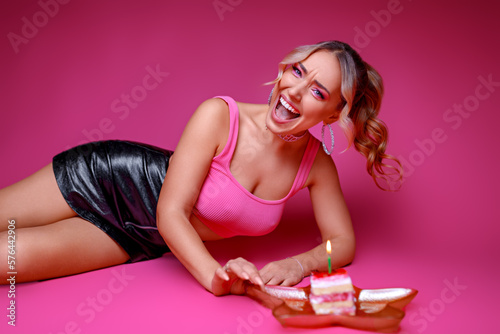 A girl with bright pink makeup lies on a pink background, with a cake and candles on a plate. Birthday.